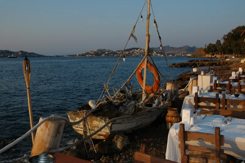 A wooden fishing boat beside a waterfront restaurant terrace on the Bodrum coast in evening light.